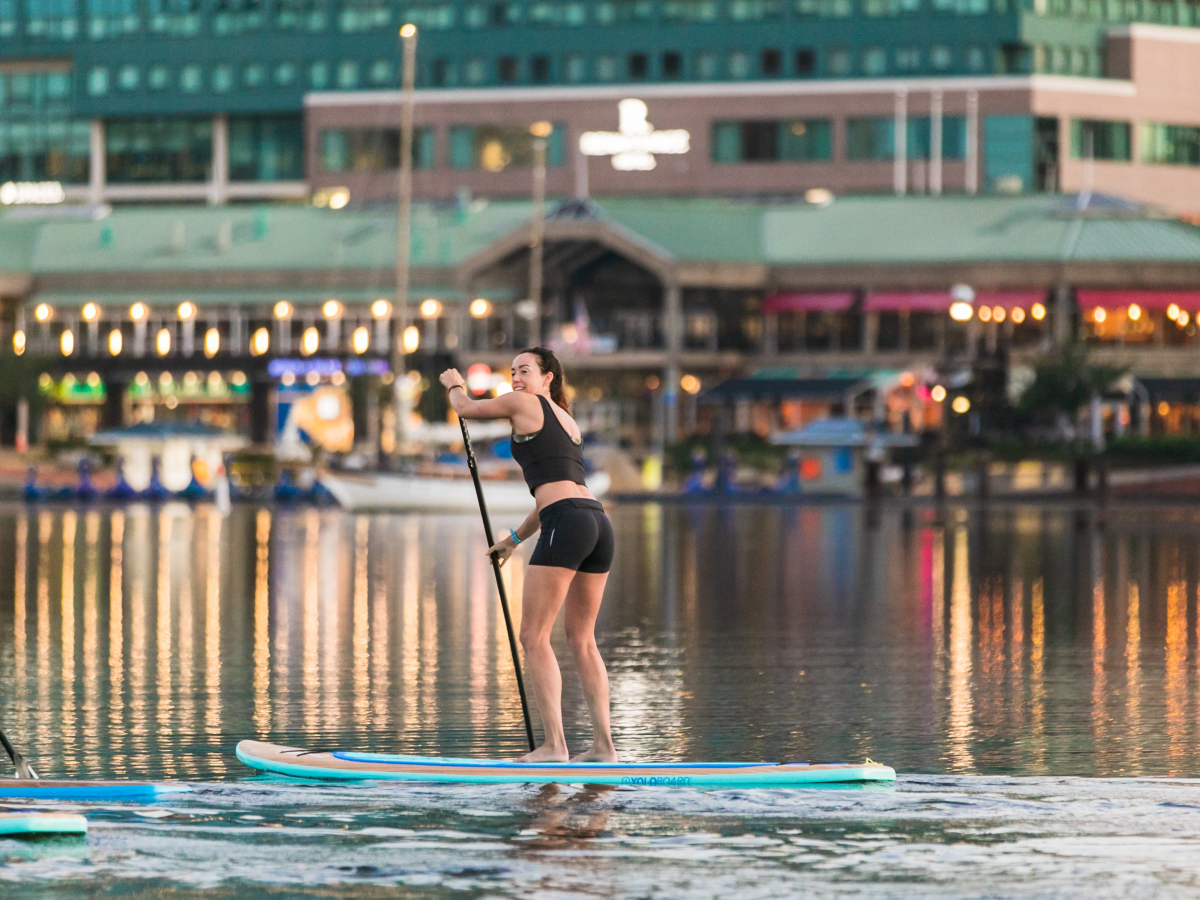 Woman paddleboarding on calm water with city lights in the background.