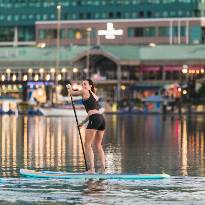 Woman paddleboarding on calm water with city lights in the background.