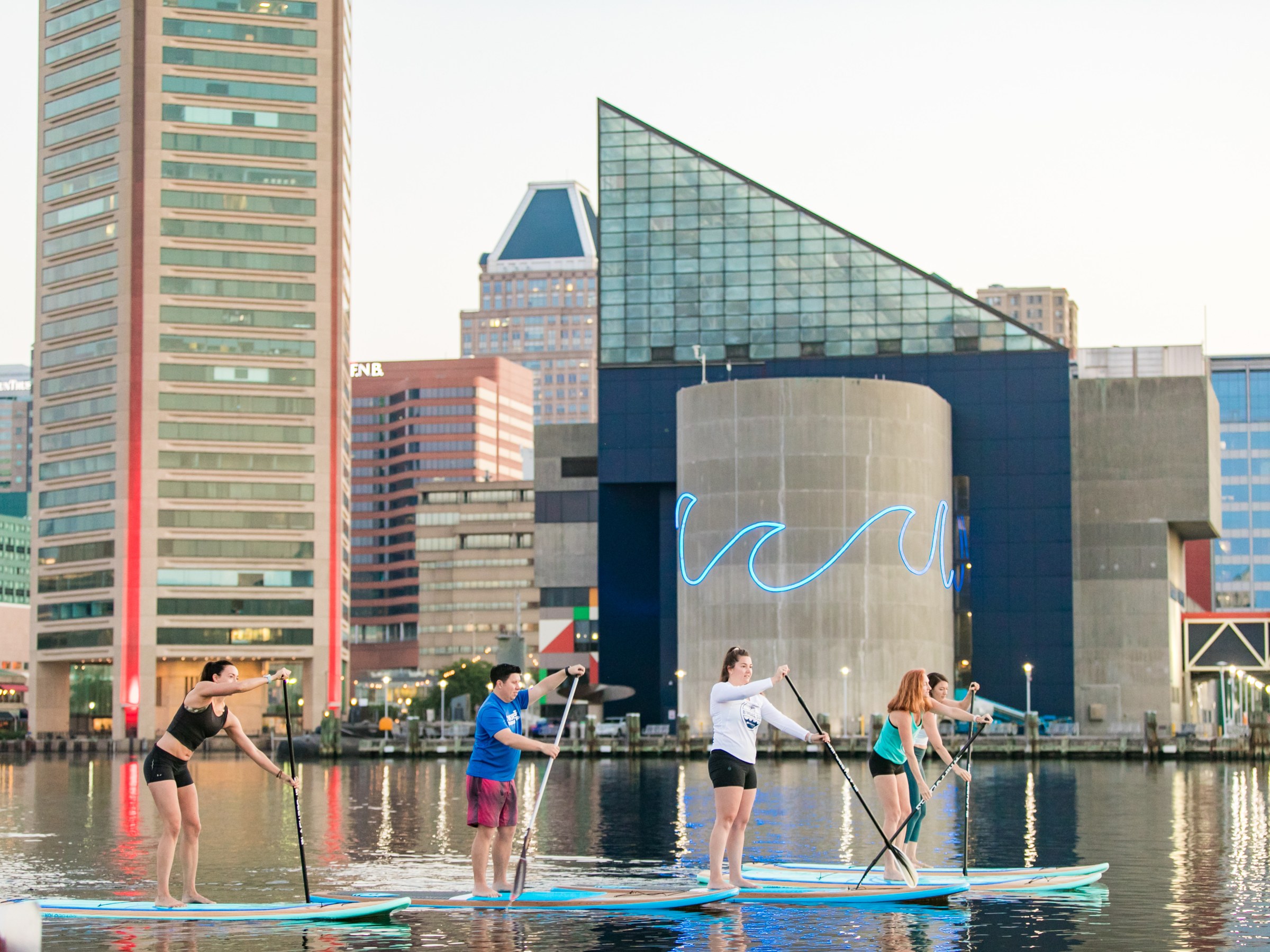 Four people paddleboarding on a calm urban waterway with modern buildings in the background.