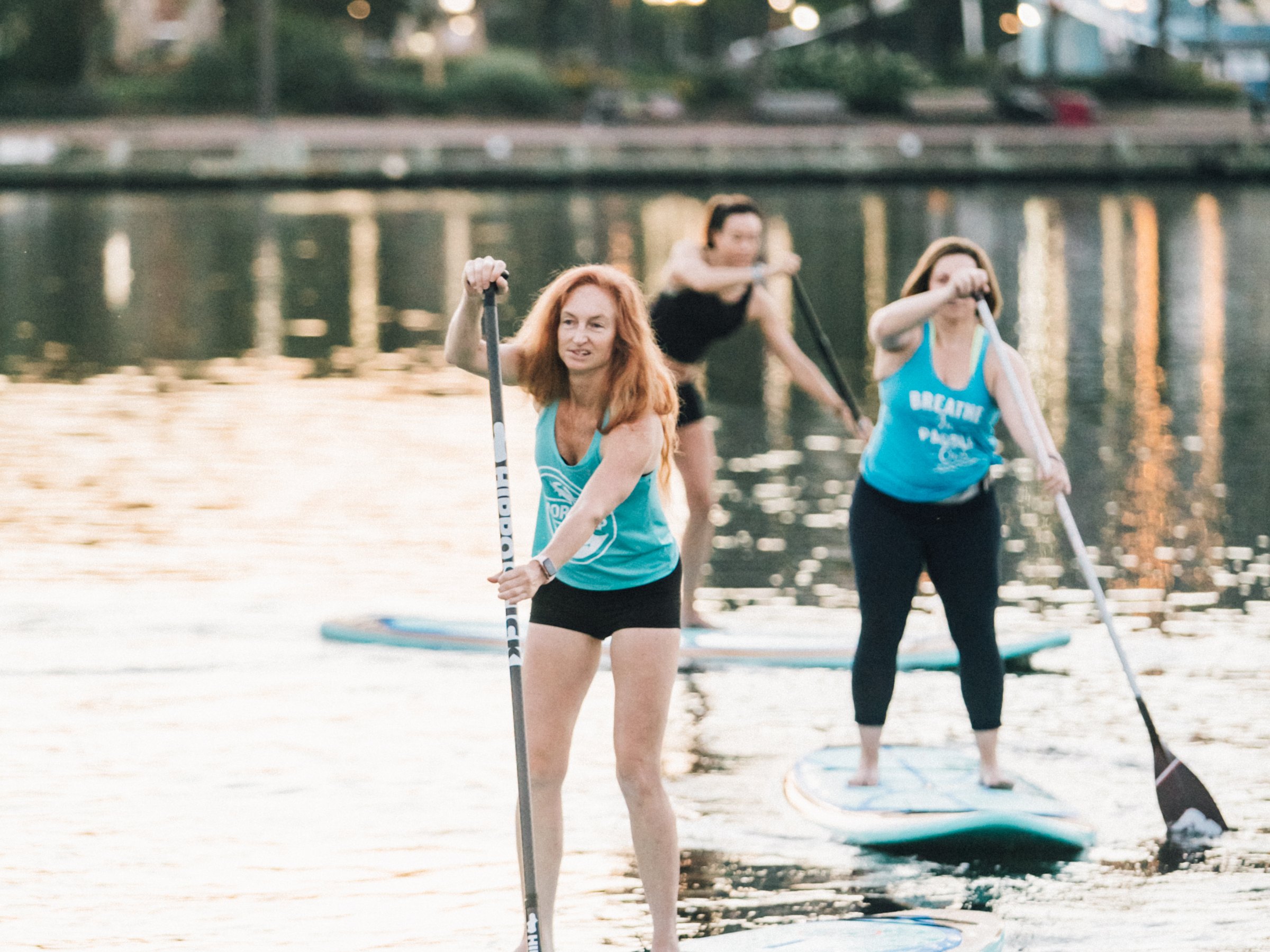 Three women stand-up paddleboarding on a calm river during sunset.