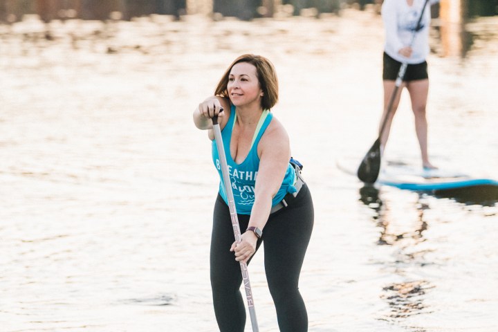 Two women paddleboarding on a calm lake with buildings in the background.