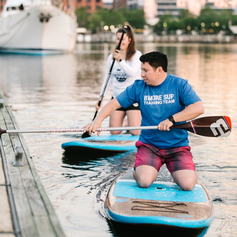 Two people paddleboarding on a calm city waterfront, one kneeling, the other standing.
