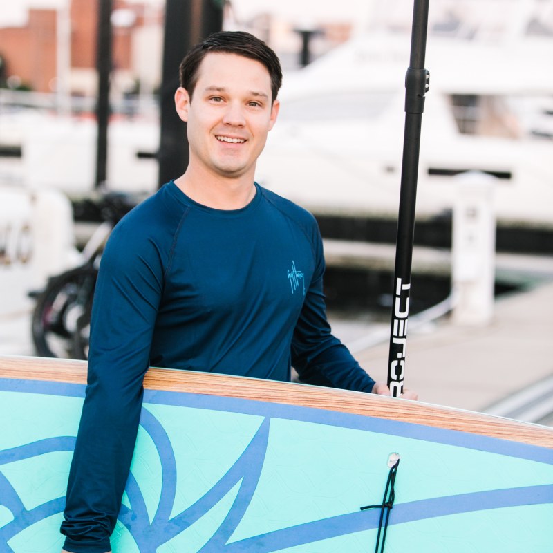 Man holding a paddleboard and paddle by a dock with boats in the background.