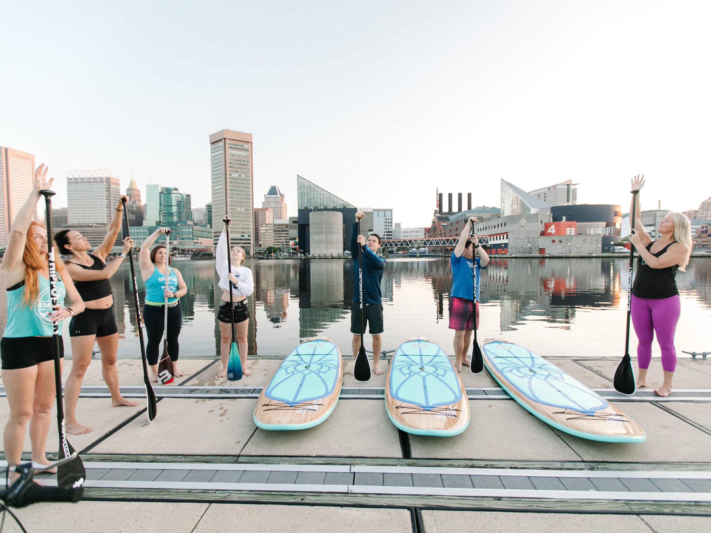 Group of people holding paddles by paddleboards near urban waterfront.