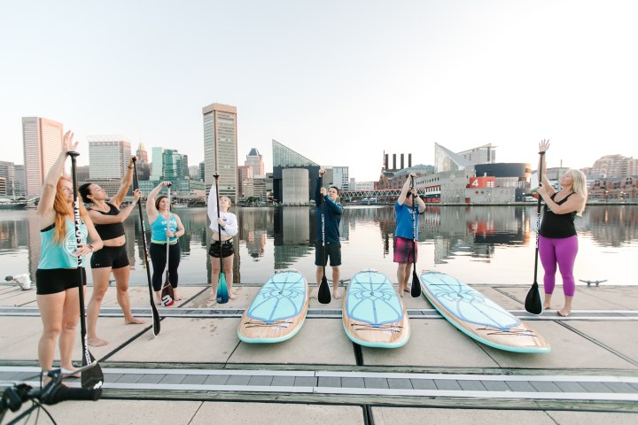 Group of people holding paddles by paddleboards near urban waterfront.