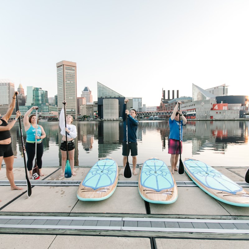 Group of people holding paddles by paddleboards near urban waterfront.
