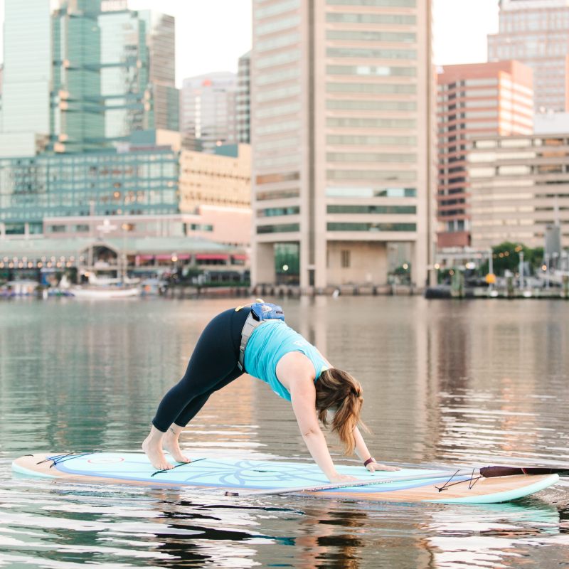 Person does yoga on a paddleboard in water, city skyline in background.