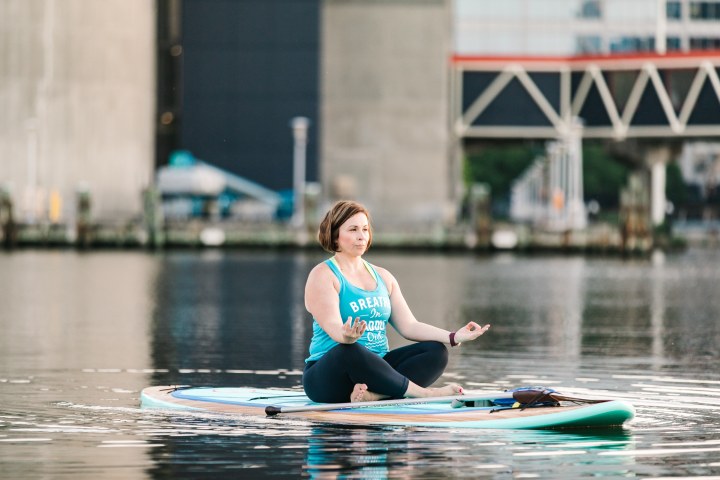 Person meditating on paddleboard in calm water near city structures.