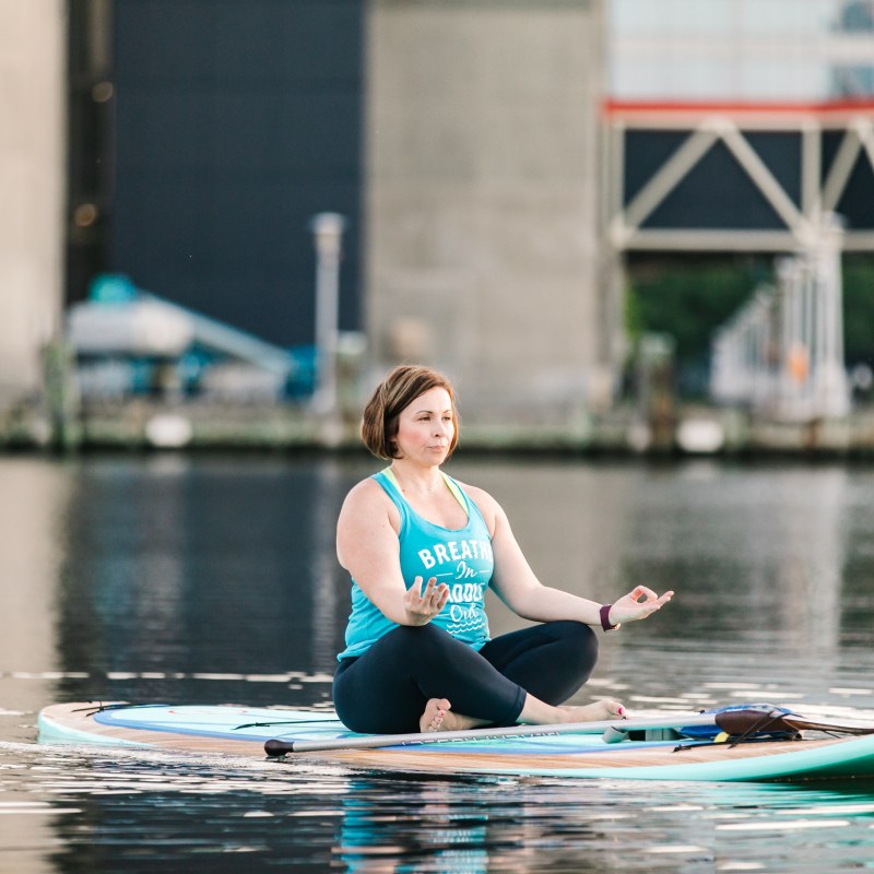 Person meditating on paddleboard in calm water near city structures.