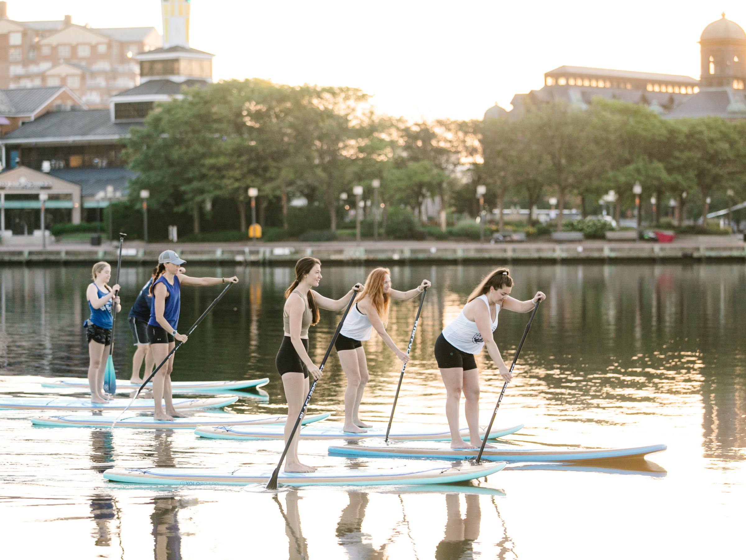 Group of people paddleboarding on a calm lake at sunset with buildings in the background.