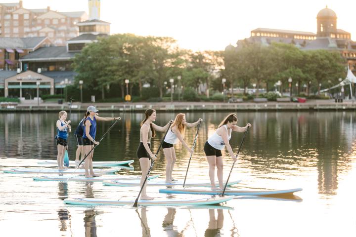 Group of people paddleboarding on a calm lake at sunset with buildings in the background.