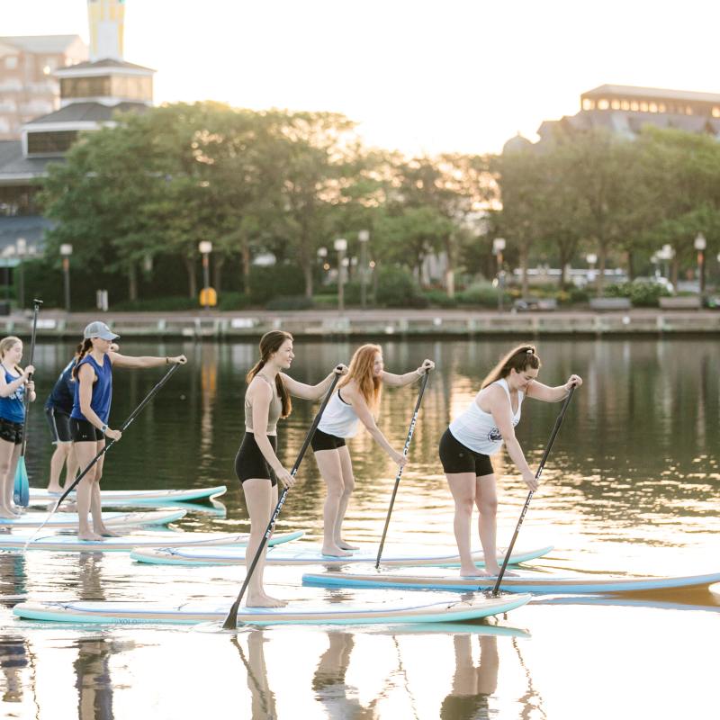Group of people paddleboarding on a calm lake at sunset with buildings in the background.