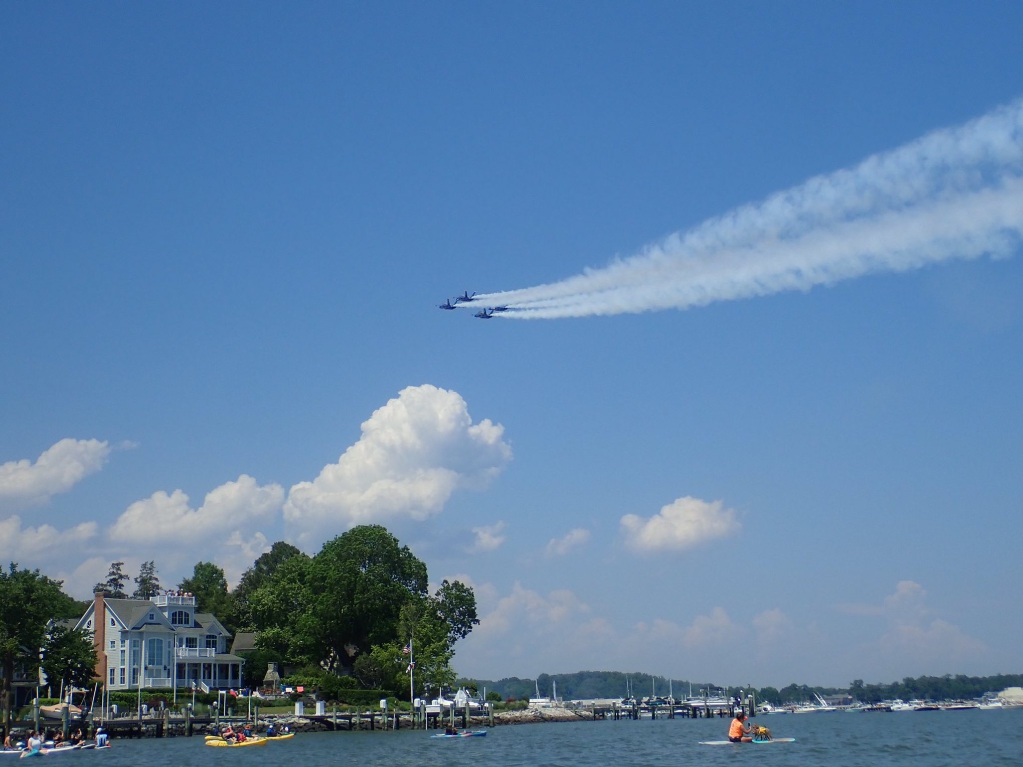 Jet formation flying over waterfront houses and kayakers on a sunny day.
