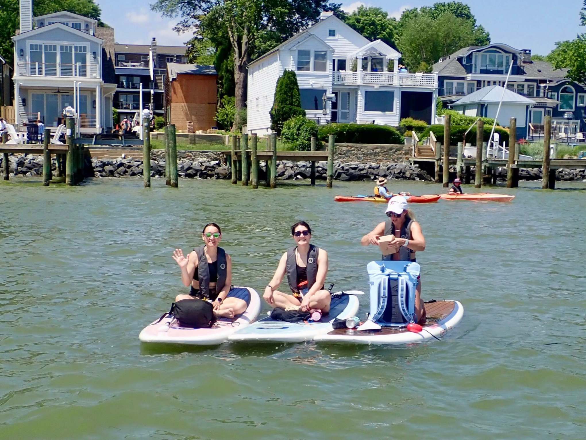 Three people on paddleboards wave near waterfront houses.