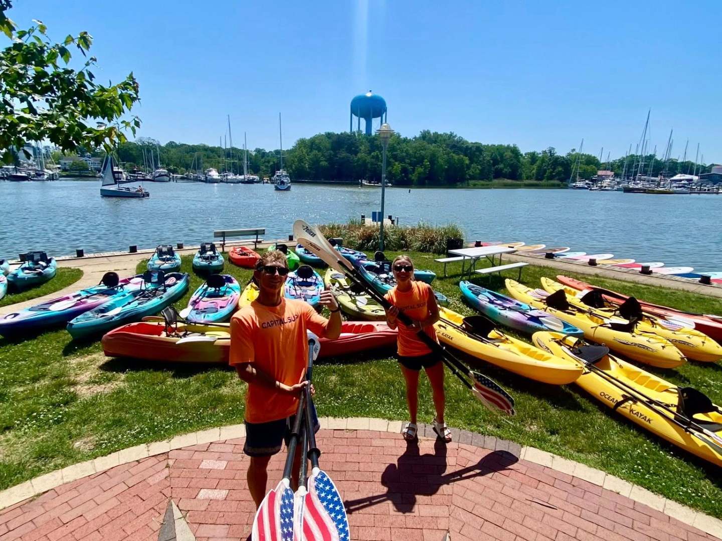Two people in orange shirts with kayaks by a lake on a sunny day.