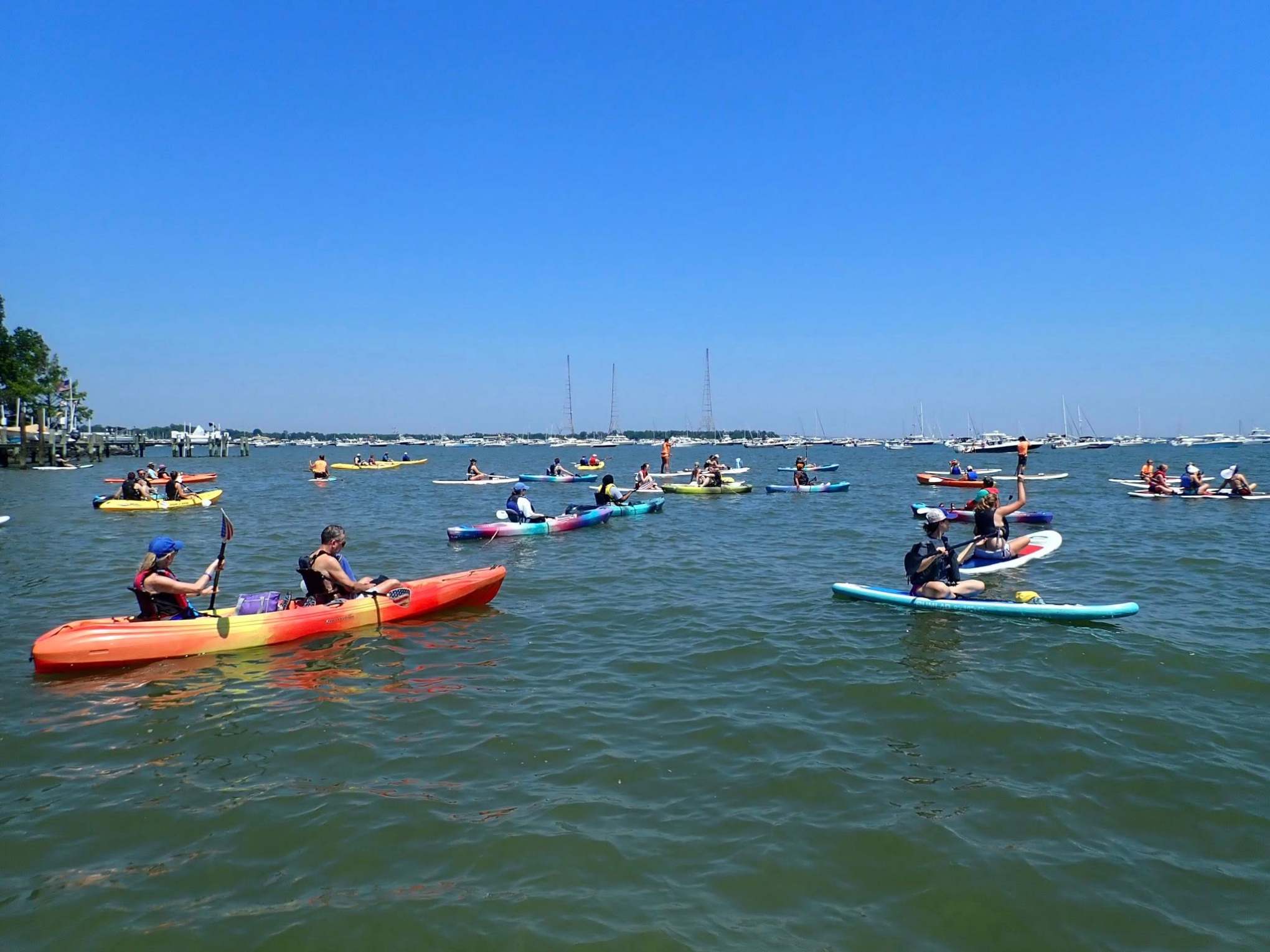 Group of people kayaking on a sunny day with boats in the background.