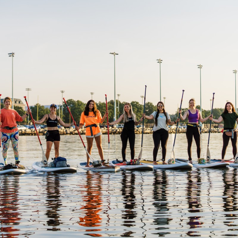 Group of people stand on paddleboards holding paddles on calm water.