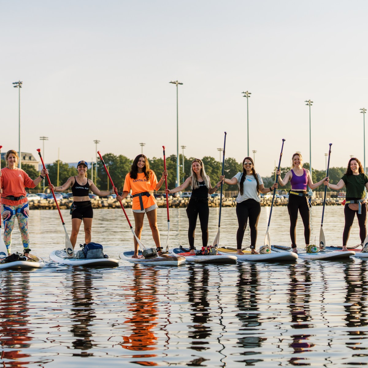 Group of people stand on paddleboards holding paddles on calm water.