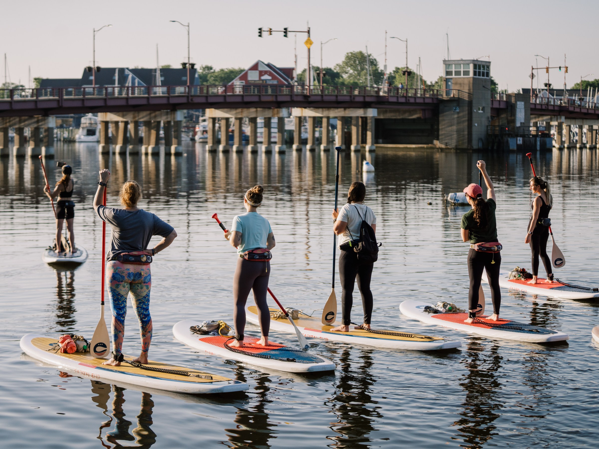 Group of people paddleboarding on a calm river near a bridge.