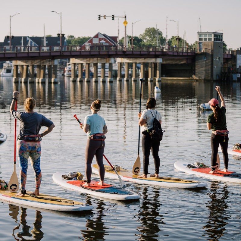 Group of people paddleboarding on a calm river near a bridge.