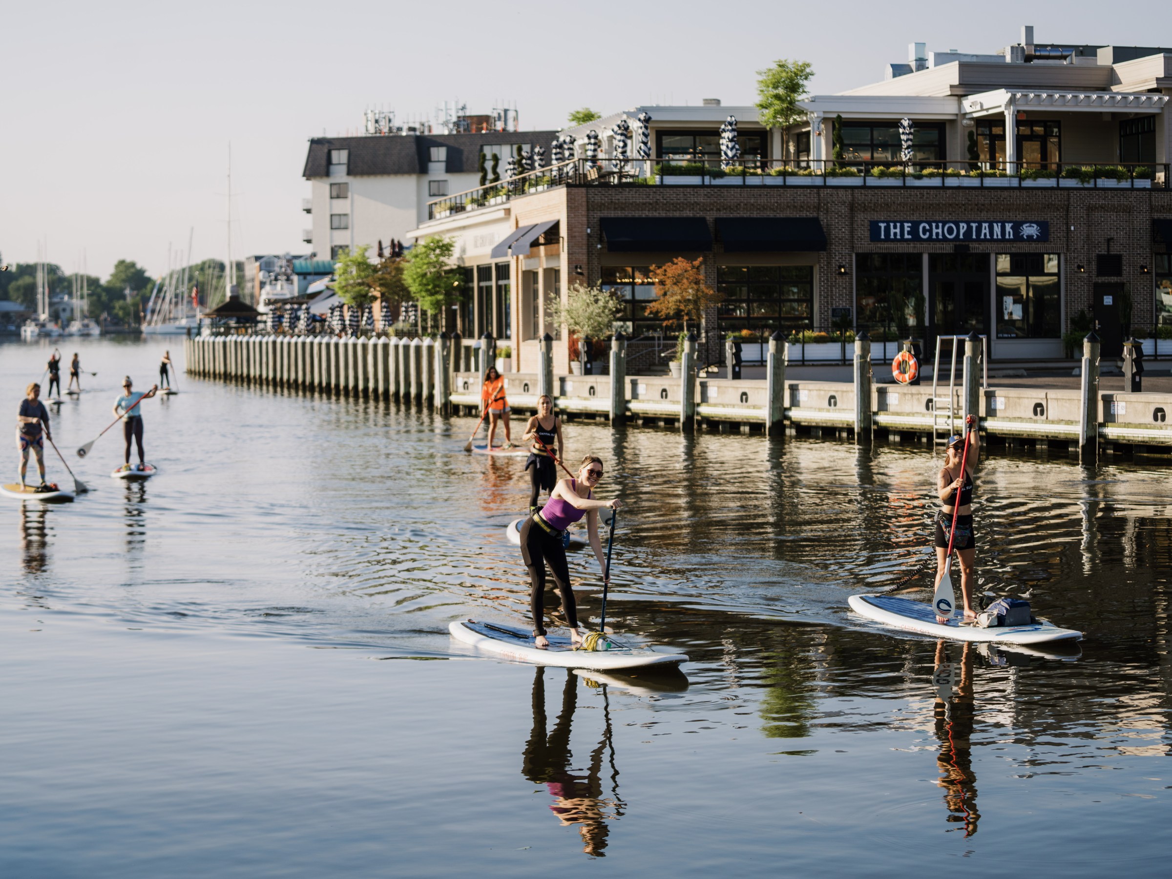 People paddleboarding on a calm waterway near a dock with buildings.