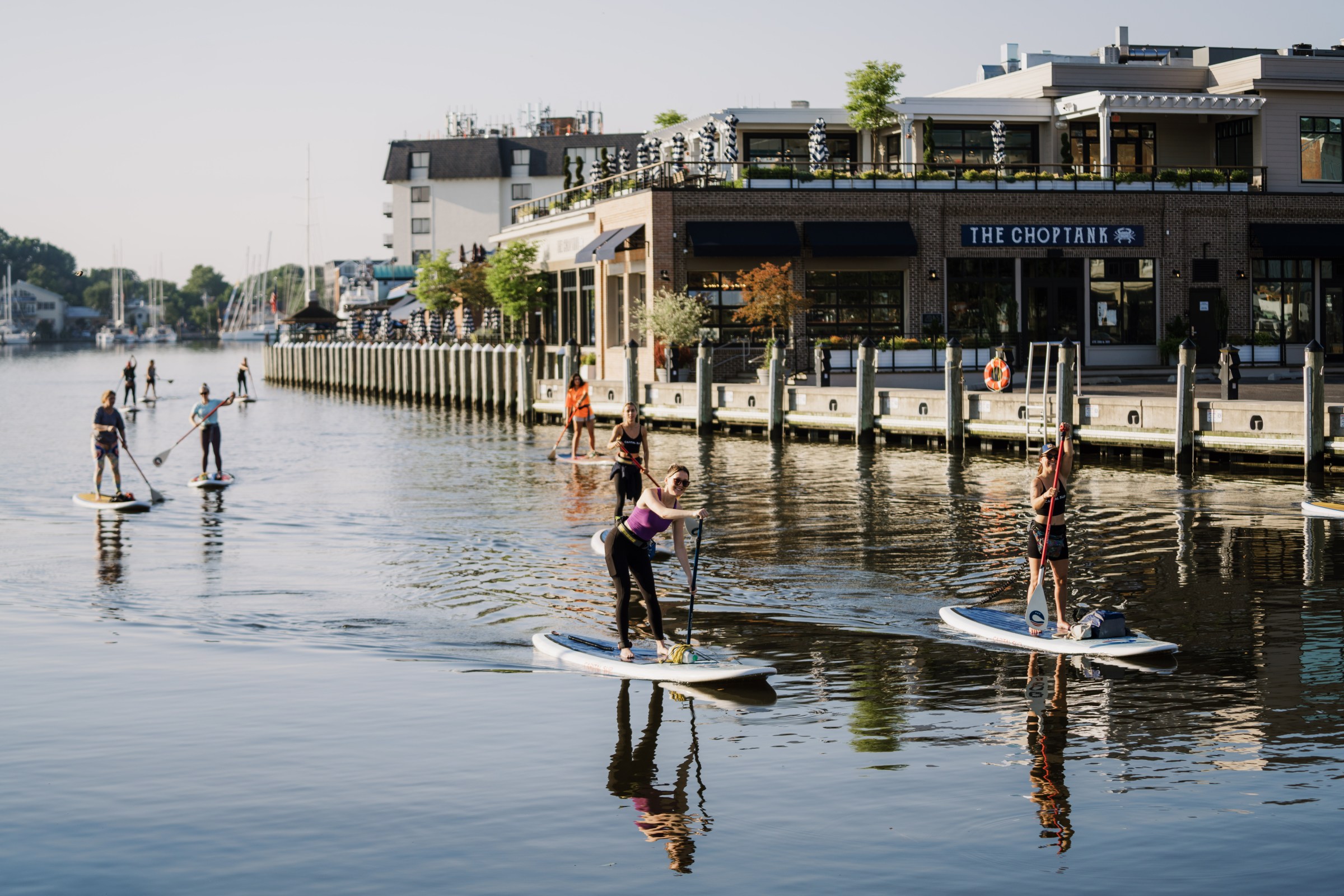 People paddleboarding on a calm waterway near a dock with buildings.