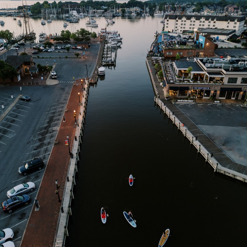 Aerial view of a canal with paddleboarders, surrounded by buildings and parked cars.