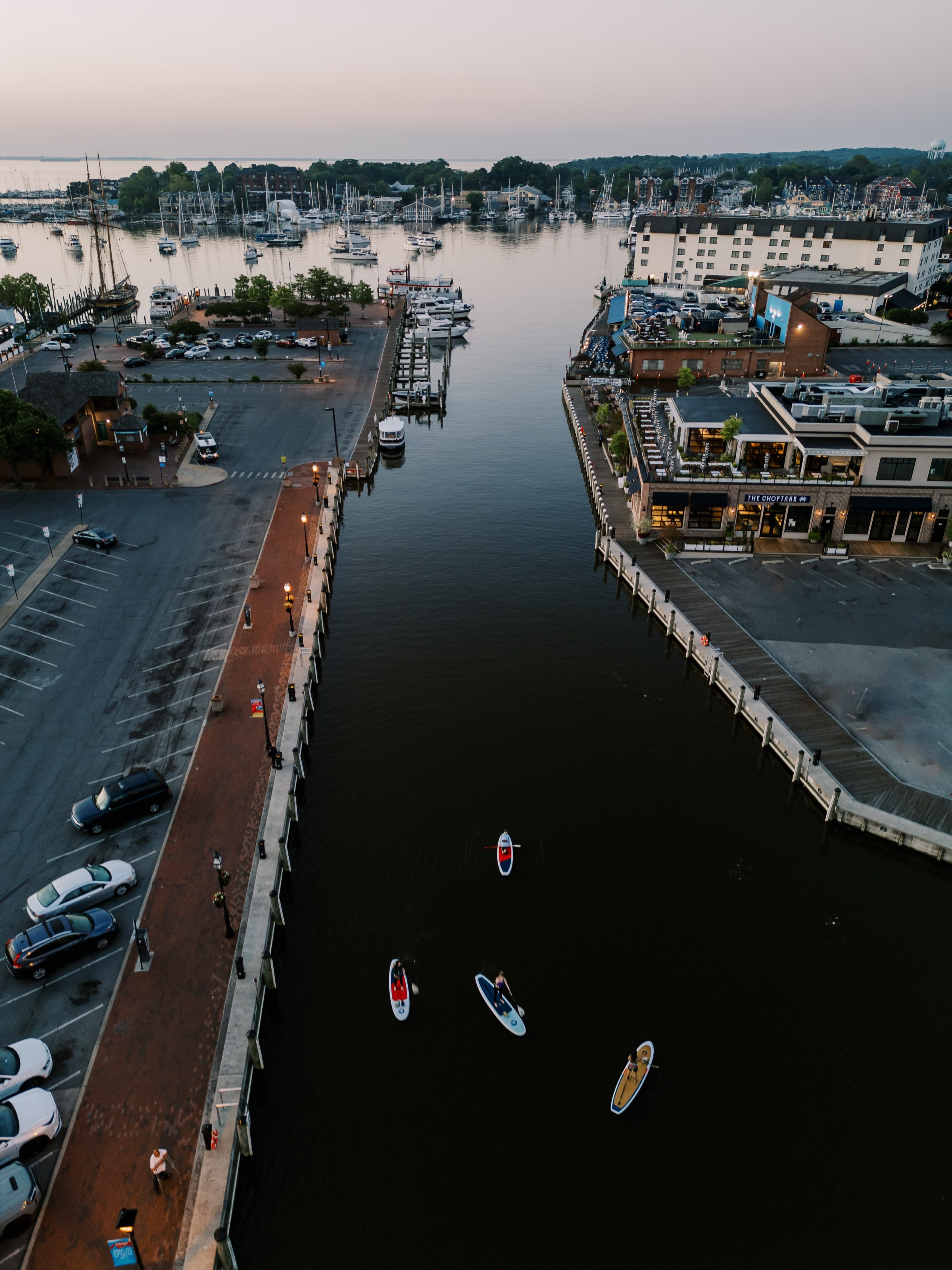 Aerial view of a canal with paddleboarders, surrounded by buildings and parked cars.