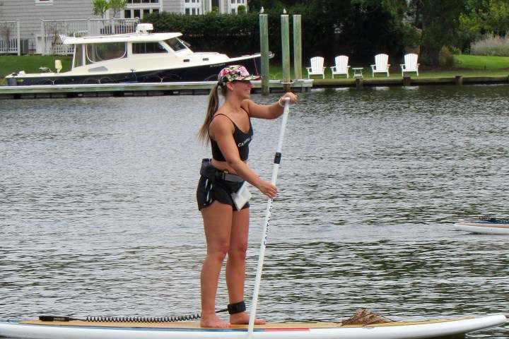 Woman paddleboarding on a river near a house and boat dock.