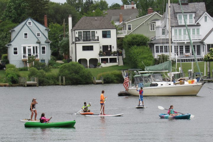People kayaking and paddleboarding on a river with houses and a boat in the background.