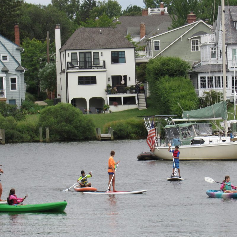 People kayaking and paddleboarding on a river with houses and a boat in the background.