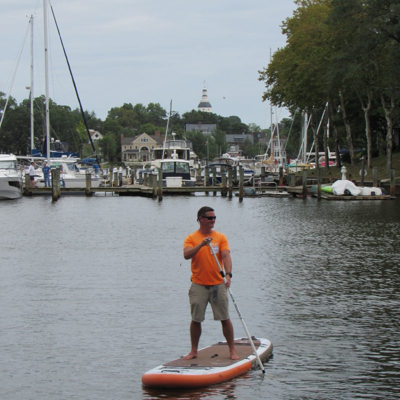 Man paddleboarding on calm water near docks with boats and buildings in background.