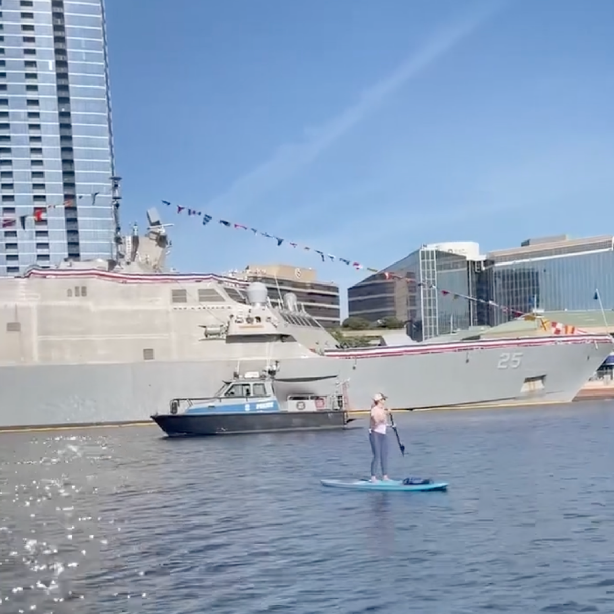 Person paddleboarding near naval ship and buildings on a sunny day.