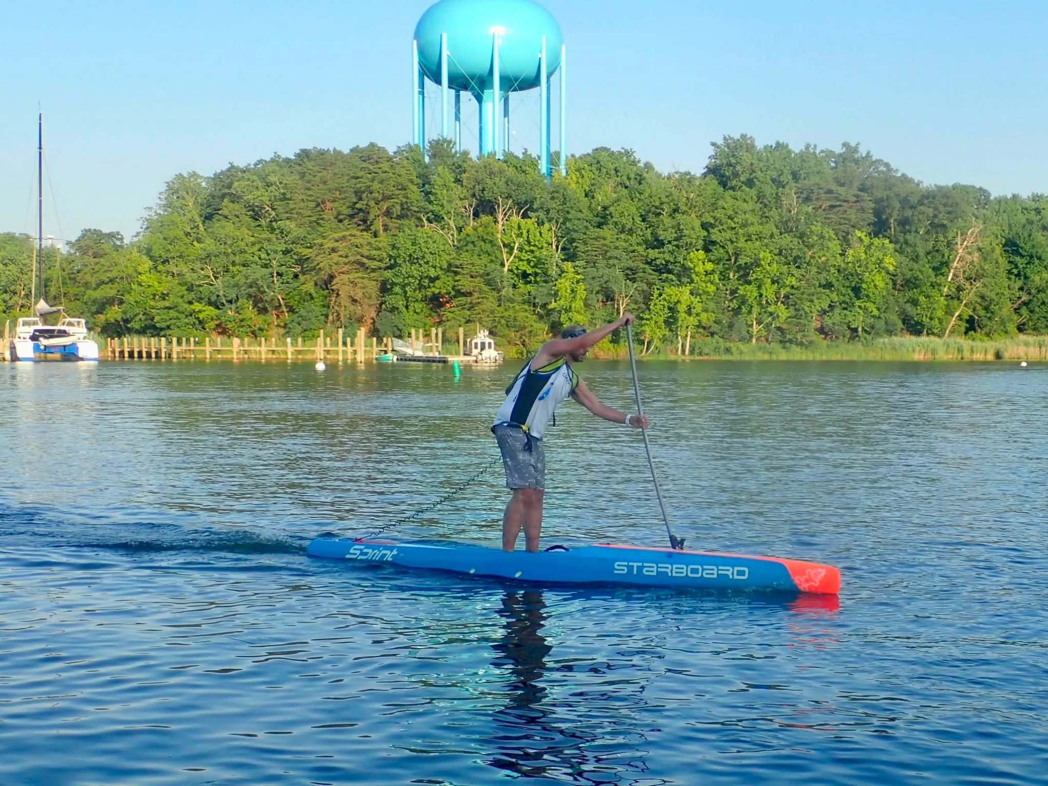 Person paddleboarding on calm water with trees and a water tower in the background.