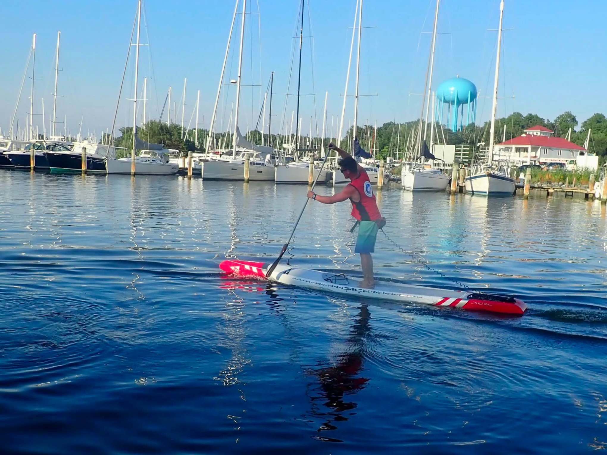 Person paddleboarding on calm marina waters with boats and a water tower in the background.