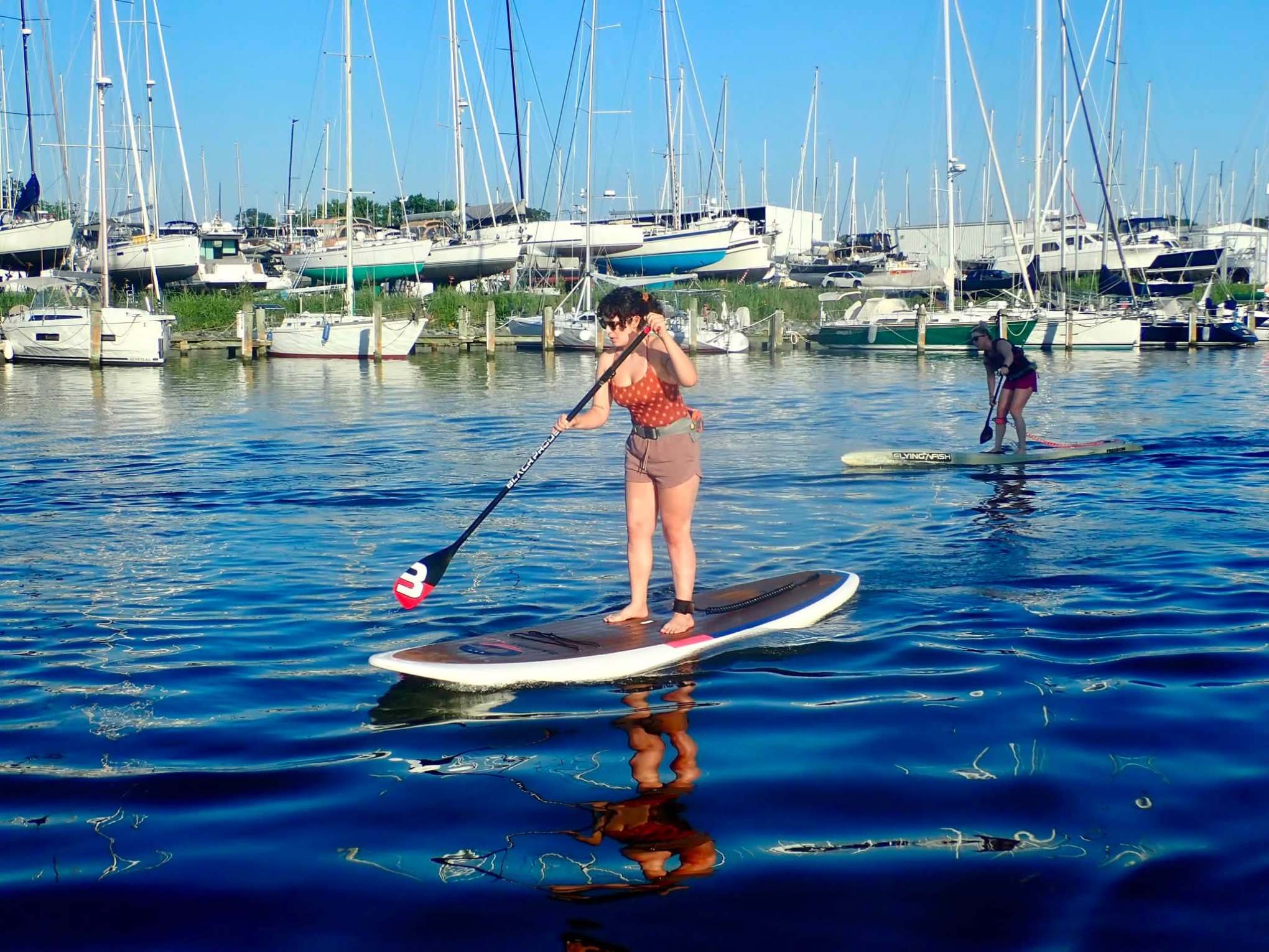 Two people paddleboarding on calm water near a marina with many boats.