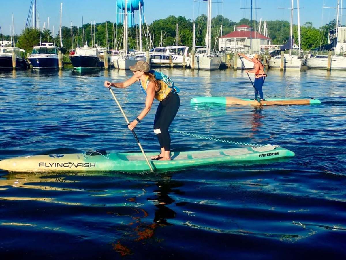 Two people paddleboarding on a marina with boats docked in the background.
