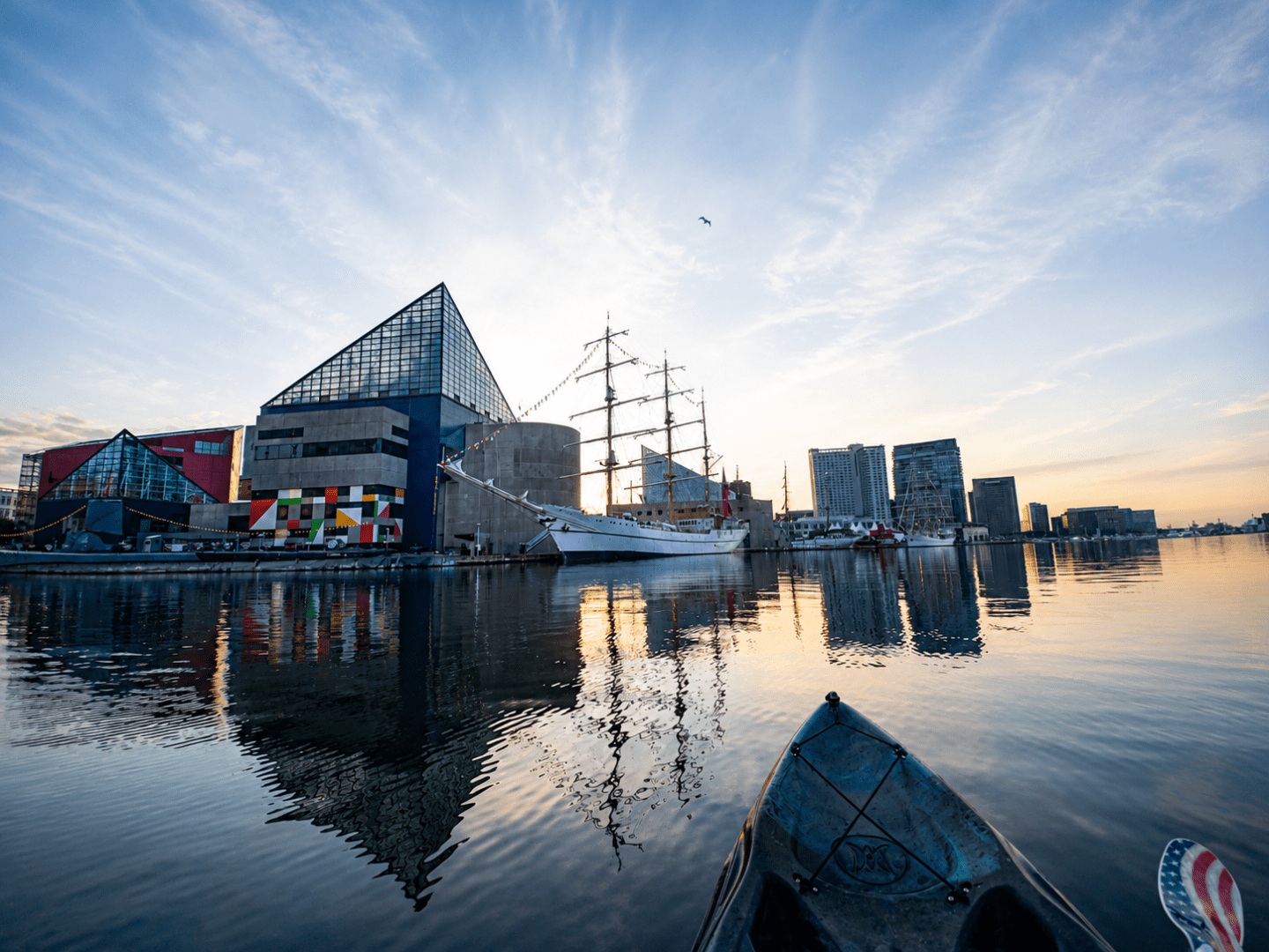 Harbor with a ship, modern buildings, and sunrise reflections on water.