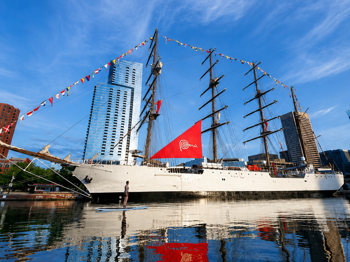 Tall ship with sails docked near modern buildings, reflection in water.