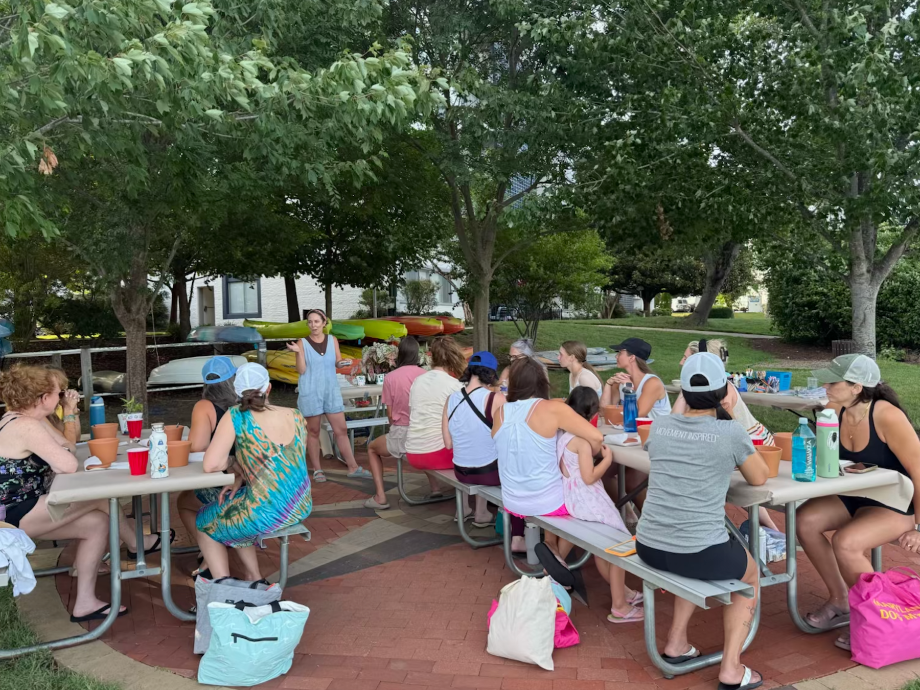 Group of people sitting at picnic tables outdoors under trees.