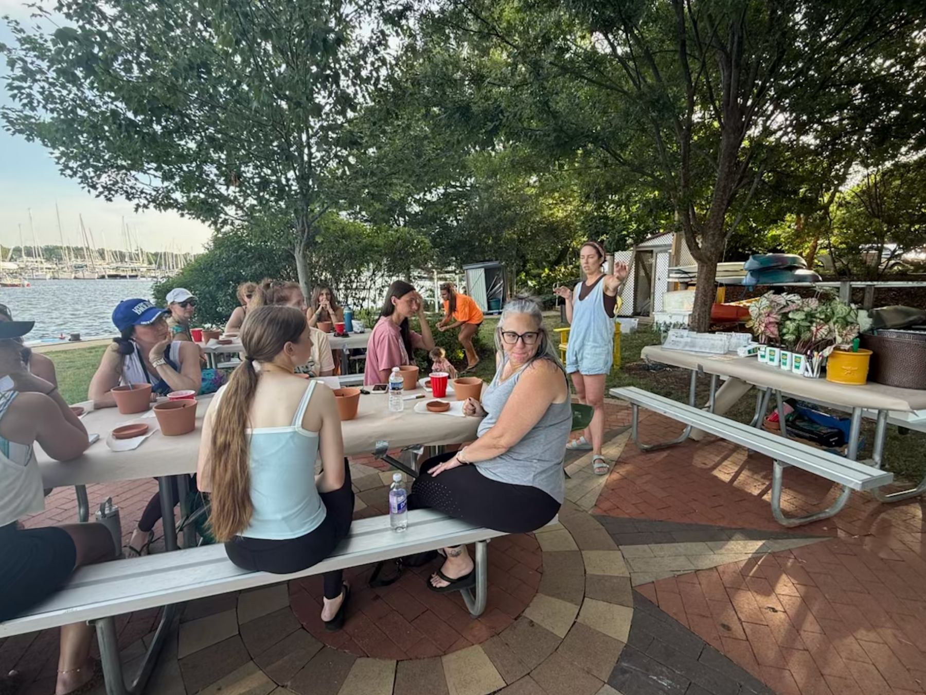 Outdoor group gathering by water, people seated at picnic tables, listening to a speaker.