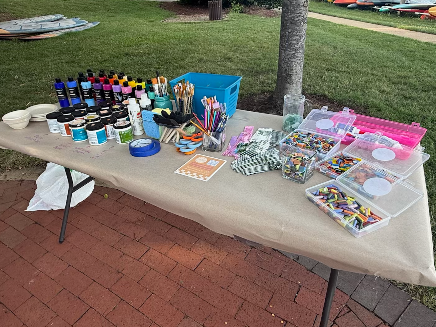 Table with art supplies like paint, brushes, and crayons on a brick sidewalk.