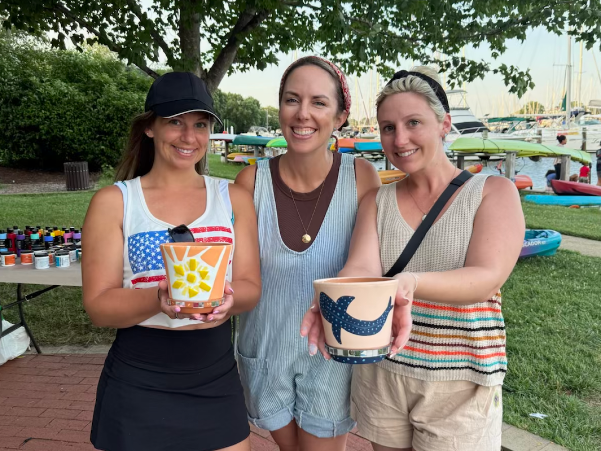 Three women smiling, holding painted flower pots outdoors.