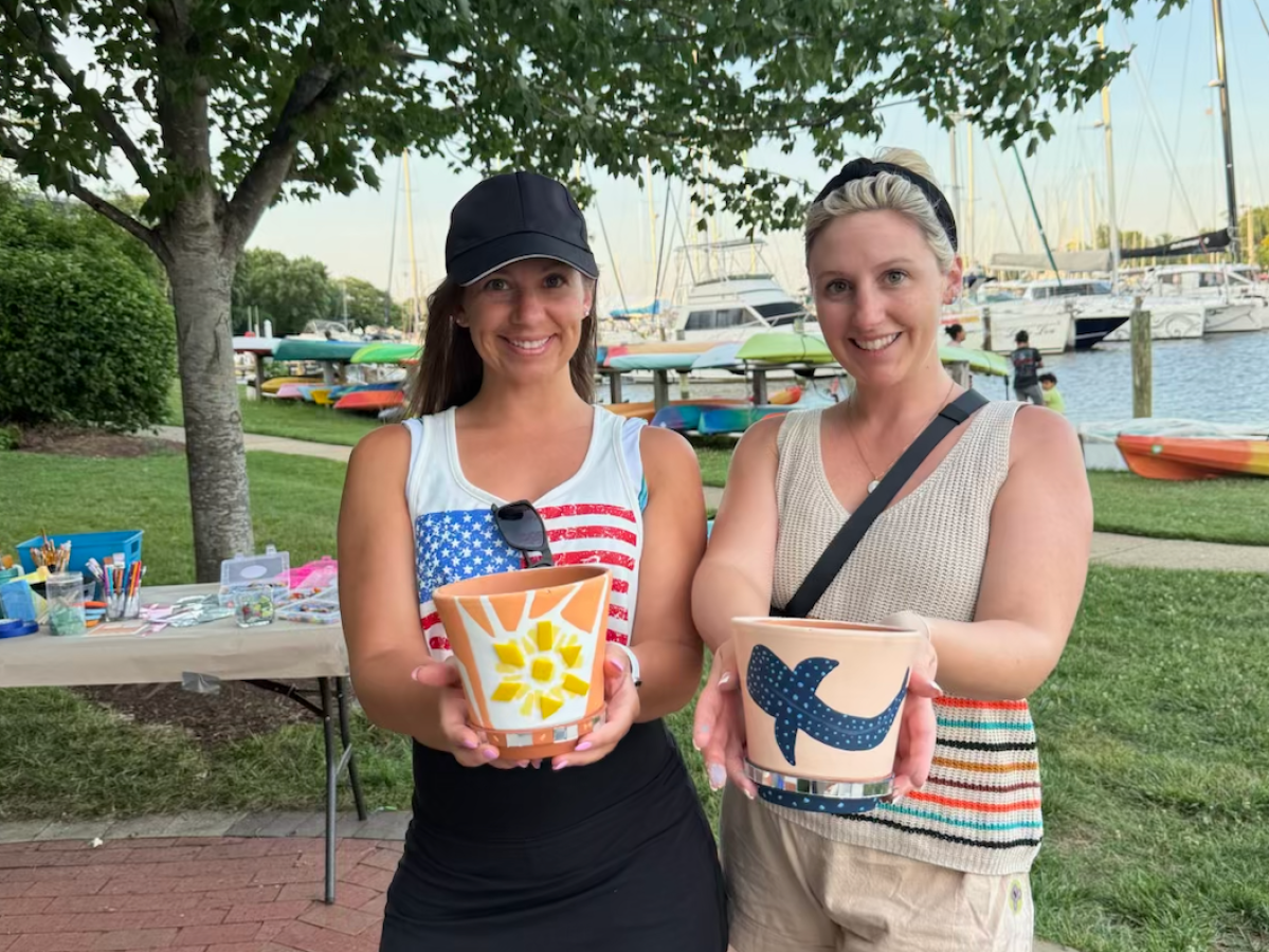 Two women outdoors holding decorated plant pots by a marina.