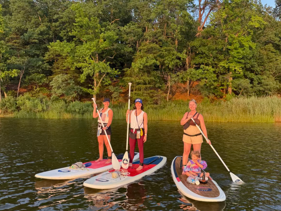 Three people stand on paddleboards with a child on a lake, trees and a water tower in the background.